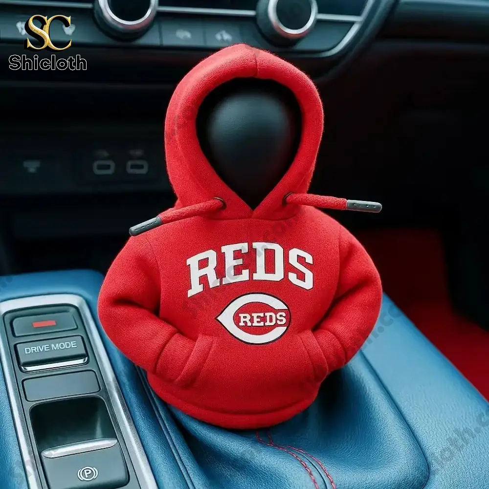 A small car gear shift cover figure wearing a red hoodie with the Reds baseball team logo inside a car interior by Shicloth!