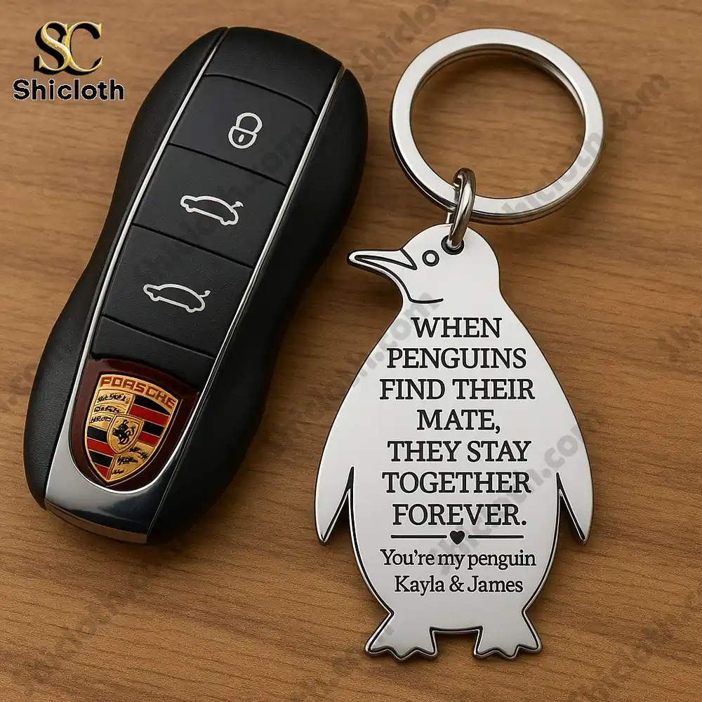 Penguin shaped keychain engraved with romantic message beside a Porsche car key on a wooden surface.