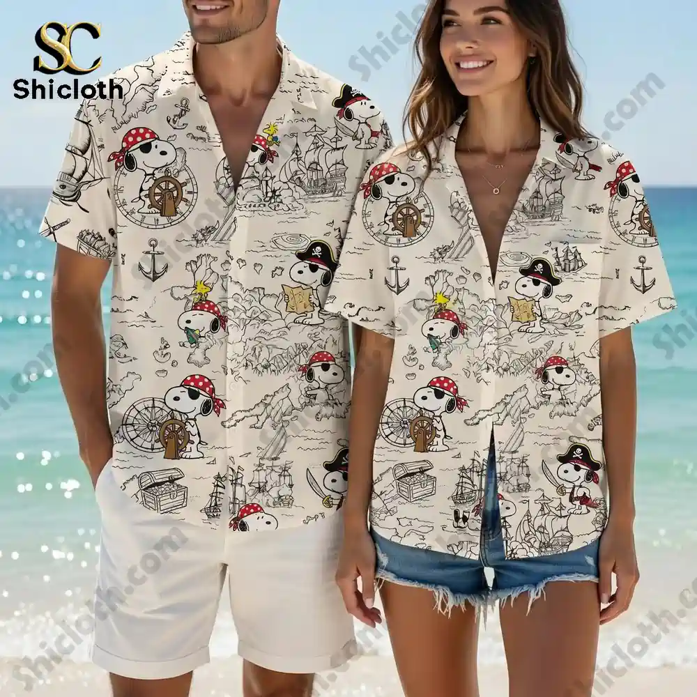 A man and a woman stand together on a sunny beach near the ocean. Both are wearing matching cream colored short sleeve button up shirts. The shirts have an all over map print with multiple illustrations of the character Snoopy dressed as a pirate along with ships and treasure chests. The man wears white shorts and the woman wears denim shorts. The water and blue sky are visible in the background.