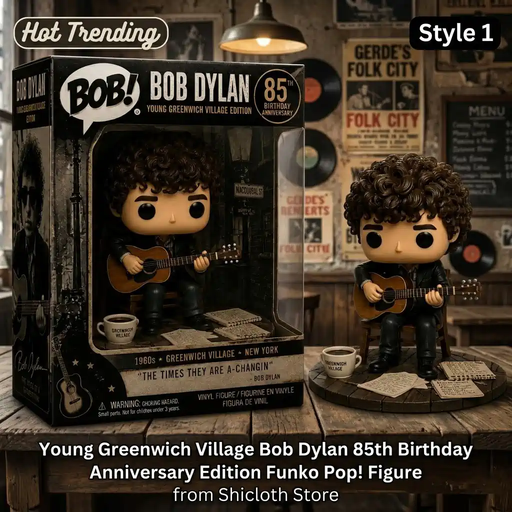 A Young Greenwich Village Bob Dylan style vinyl figure is sitting on a small chair with an acoustic guitar! It is posed next to its collector box which says 85th birthday anniversary! There is a small coffee cup and loose papers with lyrics on a weathered wooden circular base! The background is a dim room with vintage posters and a chalkboard menu!